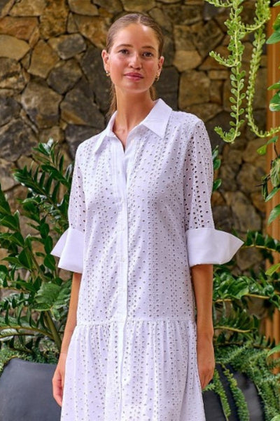 A woman wearing a white Namastai dress from Ivy Vibe Boutique, standing in a sunlit outdoor setting. The dress features a flowy silhouette and delicate details, highlighting its feminine and versatile design."